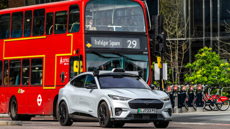 UK government brings forward self-driving pilots to spring 2026 A self-driving vehicle drives on a London street, in front of a red London bus.