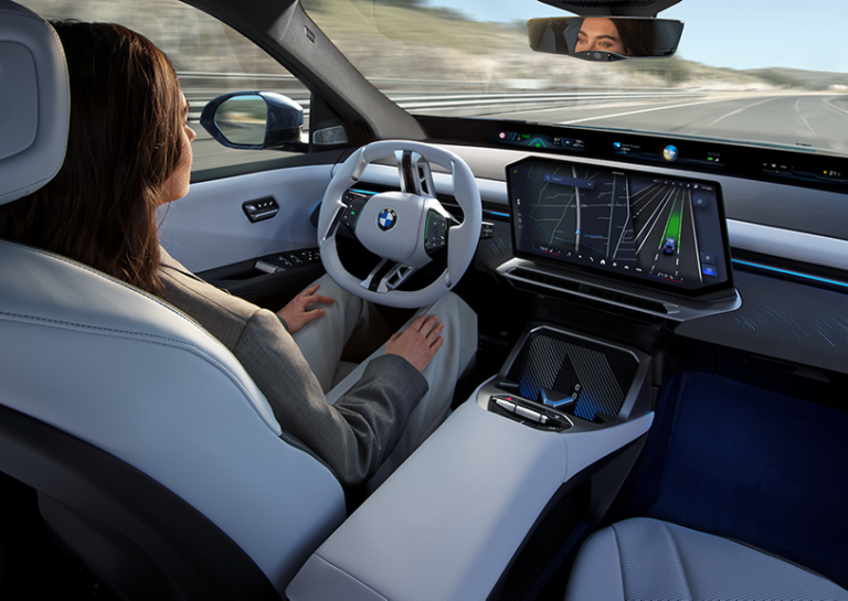A lady sits in a BMW autonomous vehicle driving down the road, with a central console display and her hands off the steering wheel