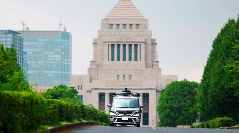 Tier IV pilot shuttle drives along an empty road with a large grey building in the background.