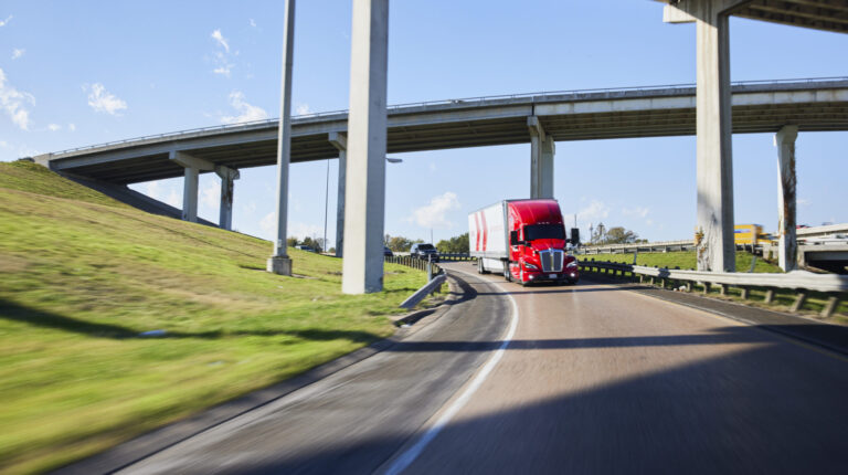 A Kodiak truck drives under a highway bridge on a bright sunny day. There are no other vehicles on the road