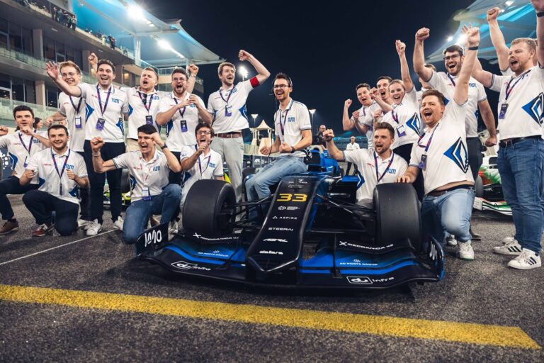 TUM team members celebrate their win at the second A2RL Grand Final with the car parked between them and the grandstand rising up on either side into the night sky