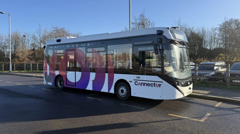 Free-to-use self-driving bus trial begins in Cambridge The Alexander Dennis Enviro100AEV electric autonomous bus driving along a quiet road on a sunny day. it has mostly white exterior with coloured writing along the side for the Connector project