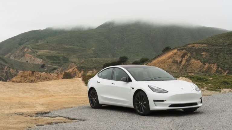 A white Tesla vehicle is parked at the edge of a hill with a misty landscape behind.