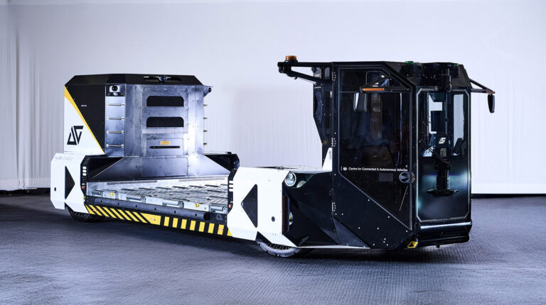 the Aurrigo automated baggage and cargo handling vehicle. It has a silver and white flat base in the middle, a larger, blakc front end and black and yellow hazard signage down the side.