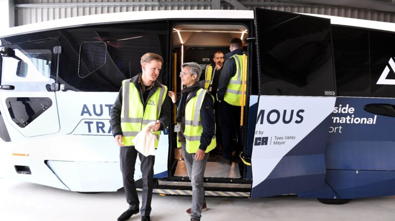 Staff in yellow high vis jackets exit a white autonomous shuttle.