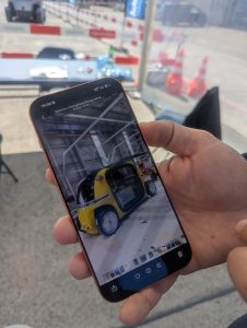 A person holds a smartphone in their hand with a yellow autonomous vehicle under test displayed on the screen. 