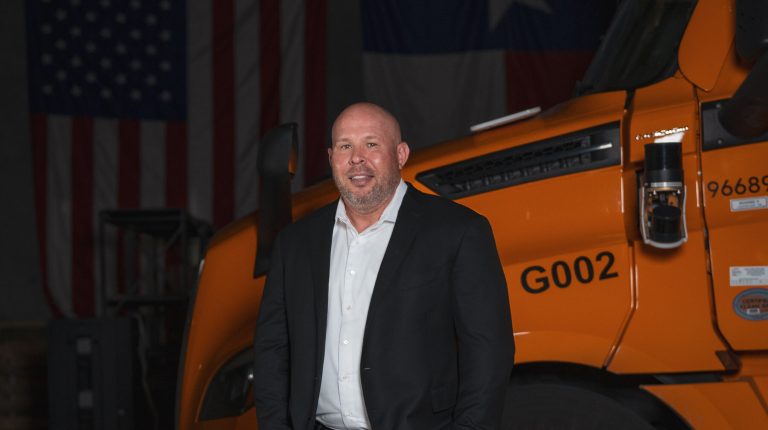 Brett Suma wears a dark suit and a white shirt, no tie. He is standing in front of an orange truck.