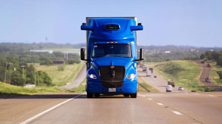 A blue second-generation Aurora driverless truck drives along a rural road on a bright sunny day.